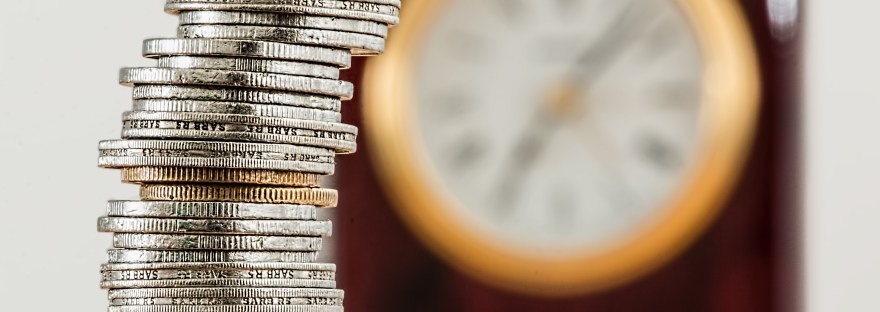 A photo of a stack of silver coins with a yellow rimmed clock in the background