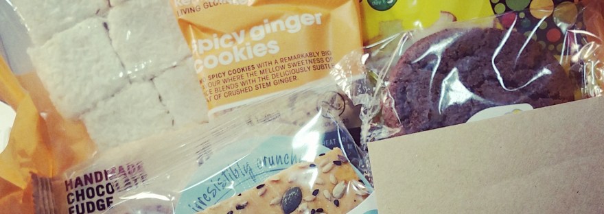 A bog of free-from treats, including: Marshmallows in a grab bag, pineapple and mango fruit crisps, 2 chocolate cookies, pack of spicy ginger cookies, some handmade fudge and a chickpea crispbread. There is a brown envelope at the front reads "Lisa" in green pen.