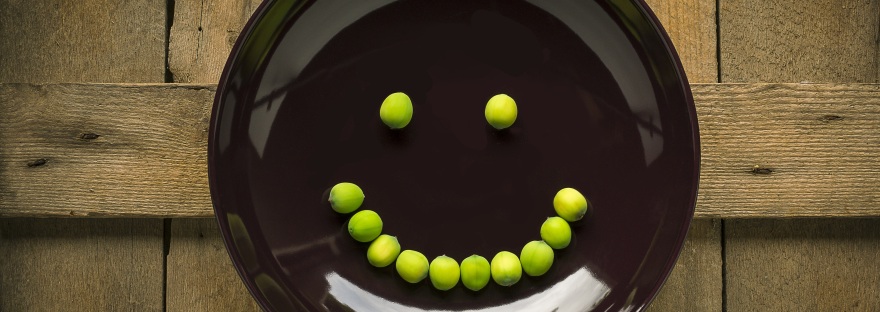 A camera down shot of a brown plate on a rough wood surface with a smiley face made of peas on the plate