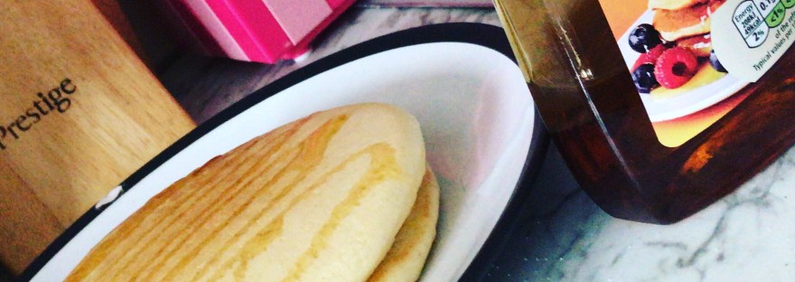A dutch angle shot of 2 fluffy pancakes on a white plate with a black rim. There is a bottle of golden syrup to the right and a knife block and Hello Kitty popcorn maker in the background. They are all on a marble surface.