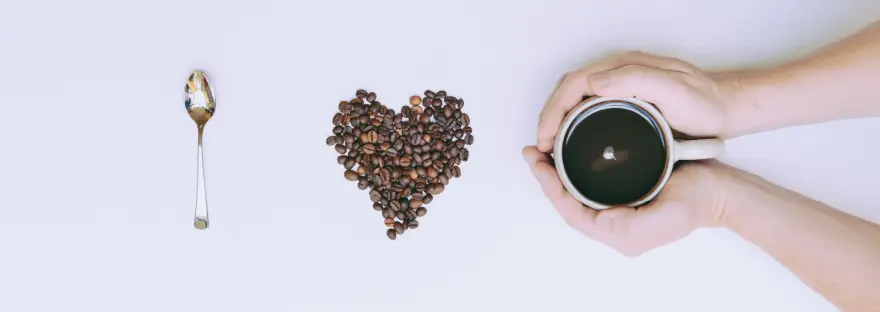 Camera Down view of a blank white canvas with: A shiny silver spoon in a vertical axis signifying the letter "I" Coffee beans in the shape of a heart to be the word "love" Last is two hands entering the frame from the right surrounding a white cup with black coffee within.